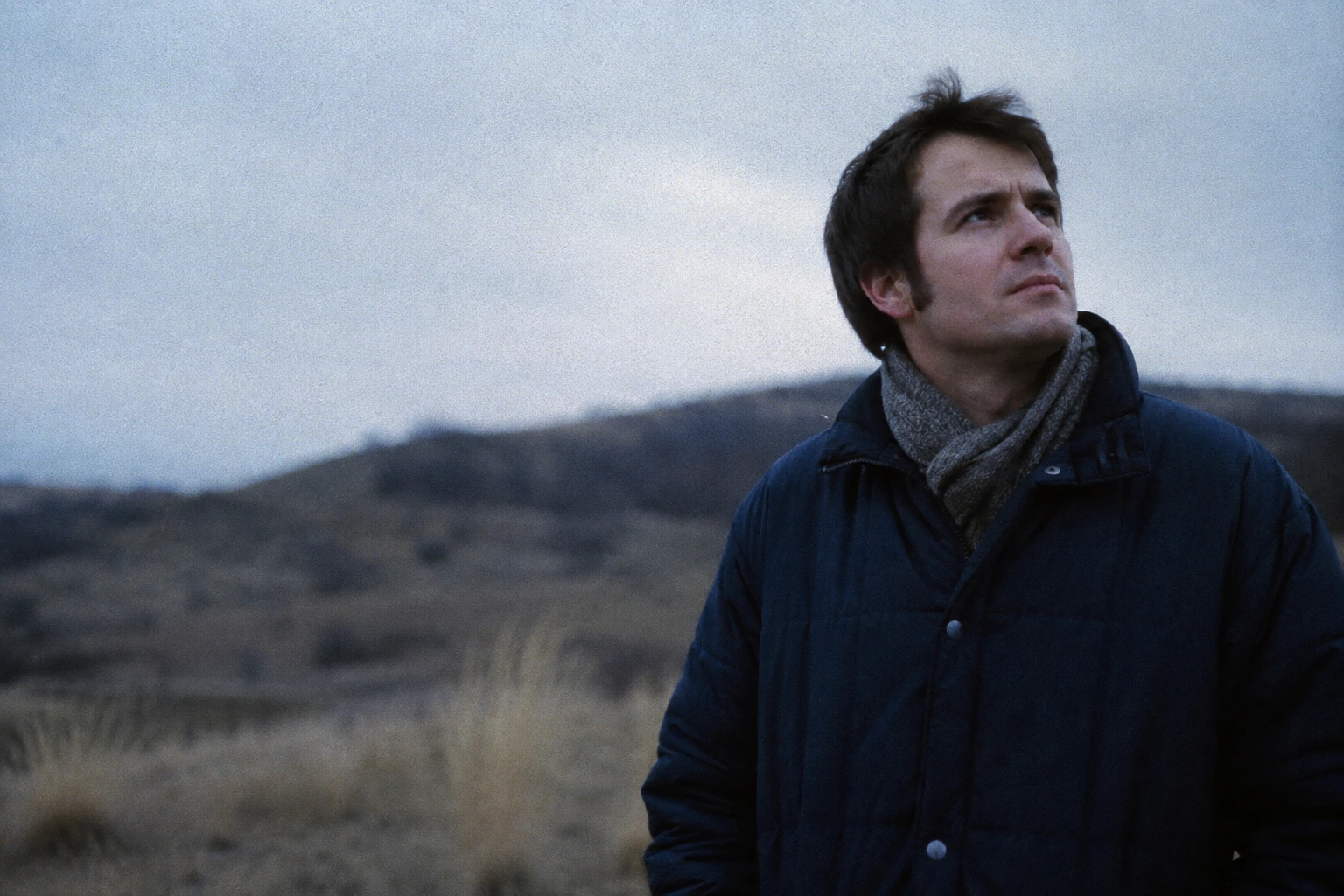 The side profile of a man in a navy parka under pale, strange light.