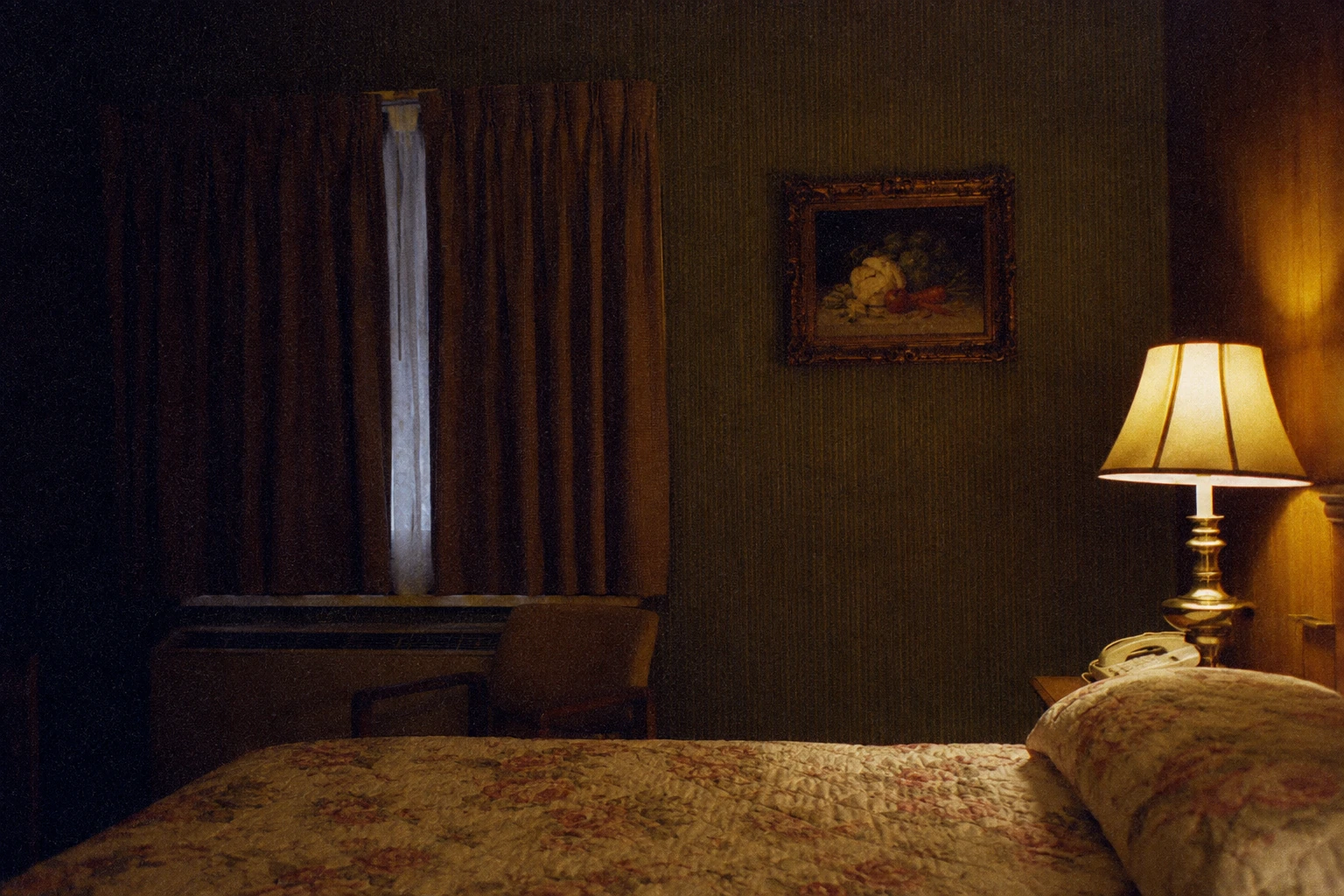 A faded hotel quilt, bedside lamp, and framed still-life in a dim 1970s room.