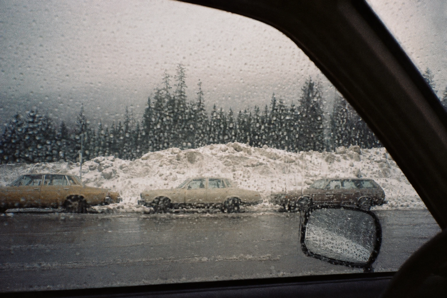 A line of 1970s cars waiting beside a tall bank of avalanche snow.