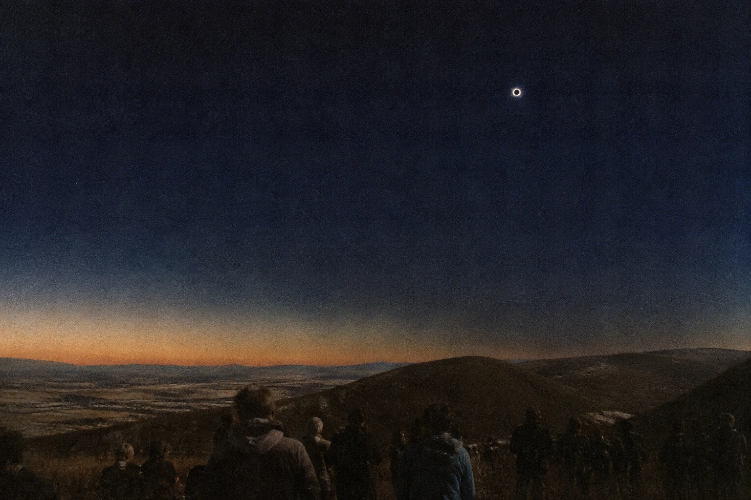A tiny white corona in an immense blue-black sky above low hills.