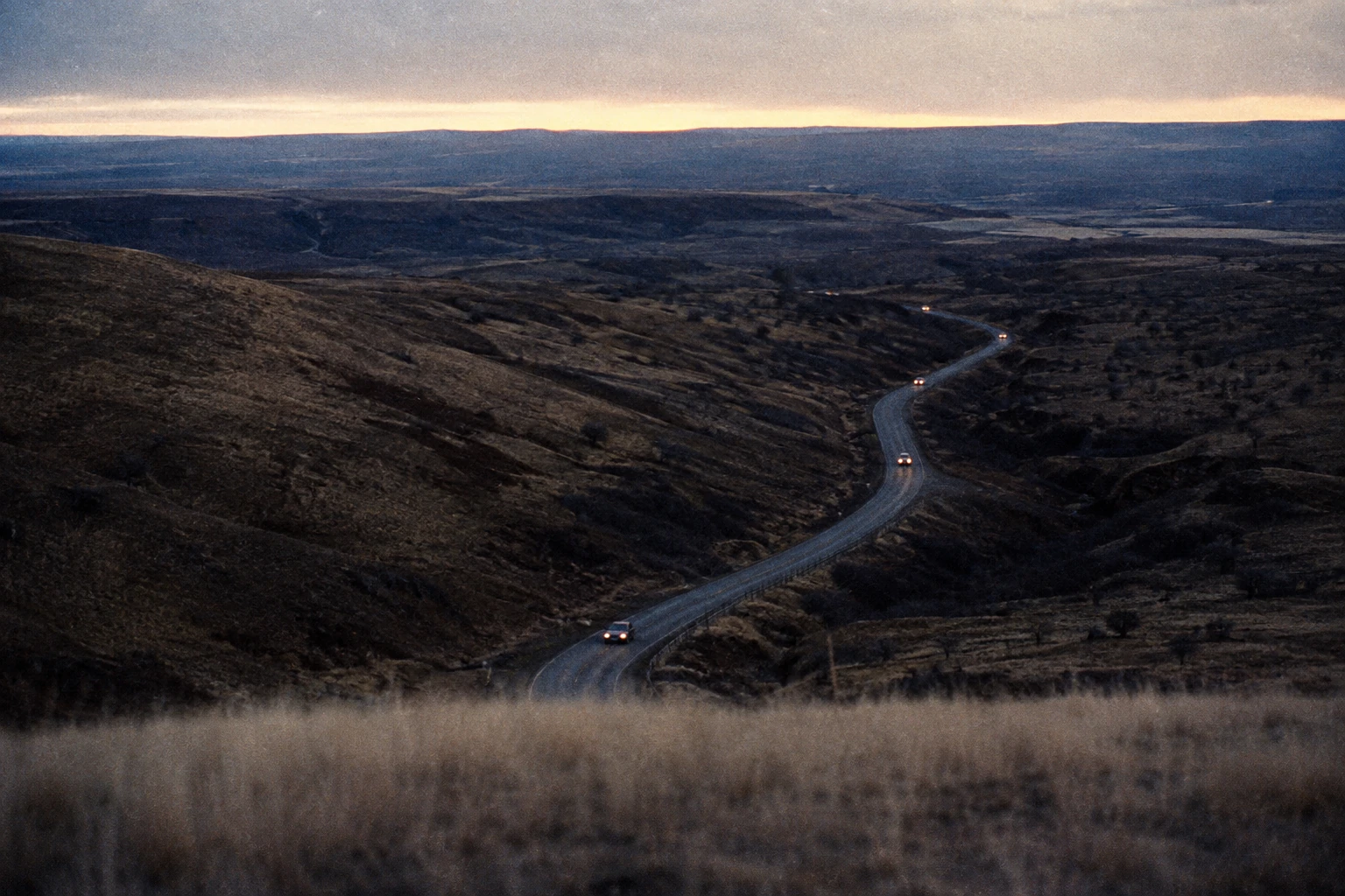 A few 1970s cars with headlights on a narrow road below a hill.